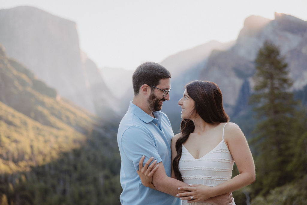 A couple embraces in a scenic mountain landscape with sunlight illuminating the background for their Yosemite national park engagement photos