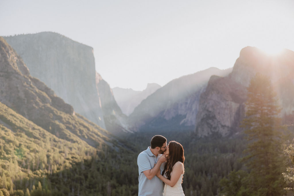 A couple embraces in front of a scenic mountain landscape with sunlight streaming over the peaks.