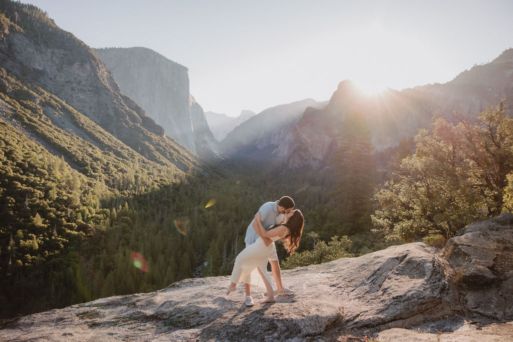 Couple embracing on a rocky overlook with a sunlit valley and mountains in the background.