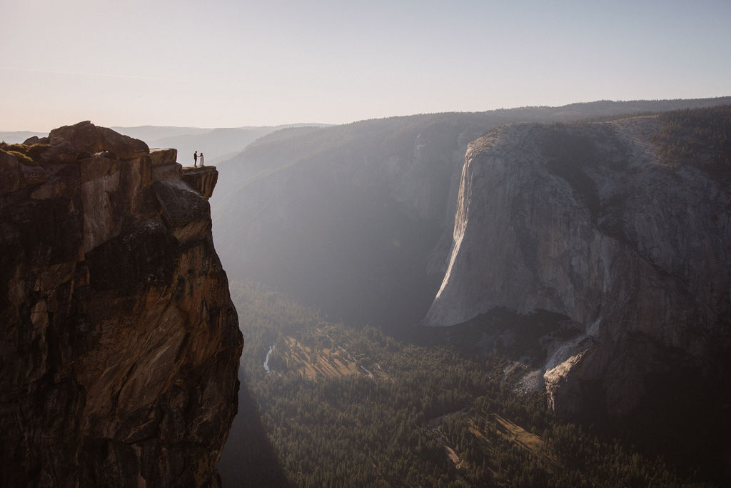 A couple dressed in wedding attire stands on a mountain ledge overlooking a vast rocky landscape with a prominent peak in the background. Best views in yosemite national park