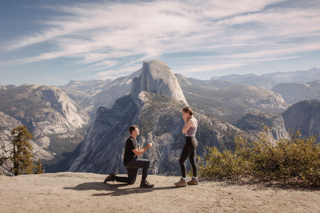 A person kneels in a proposal pose in front of another person on a mountain overlook with a large rock formation in the background under a partly cloudy sky. | How to Plan a Surprise Proposal at Yosemite National Park