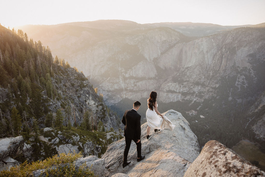 A couple dressed in wedding attire stands on a mountain ledge overlooking a vast rocky landscape with a prominent peak in the background. Best views in yosemite national park