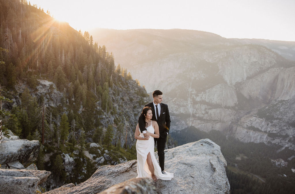 A couple dressed in wedding attire stands on a mountain ledge overlooking a vast rocky landscape with a prominent peak in the background. Best views in yosemite national park