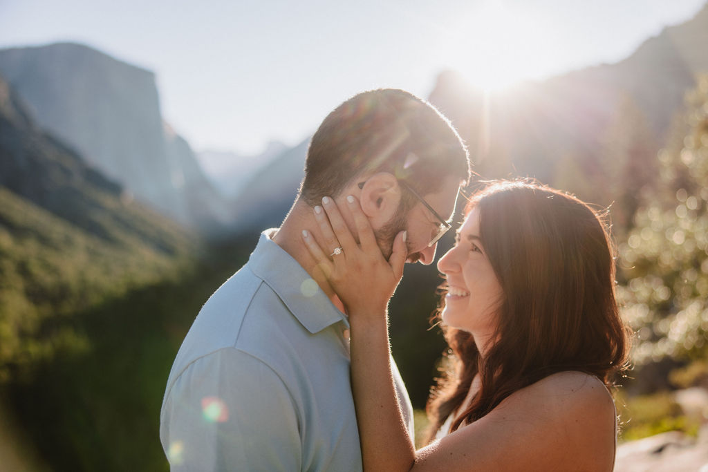 woman holds mans face and smiles while the sun peaks through them for their Yosemite National Park engagement photos