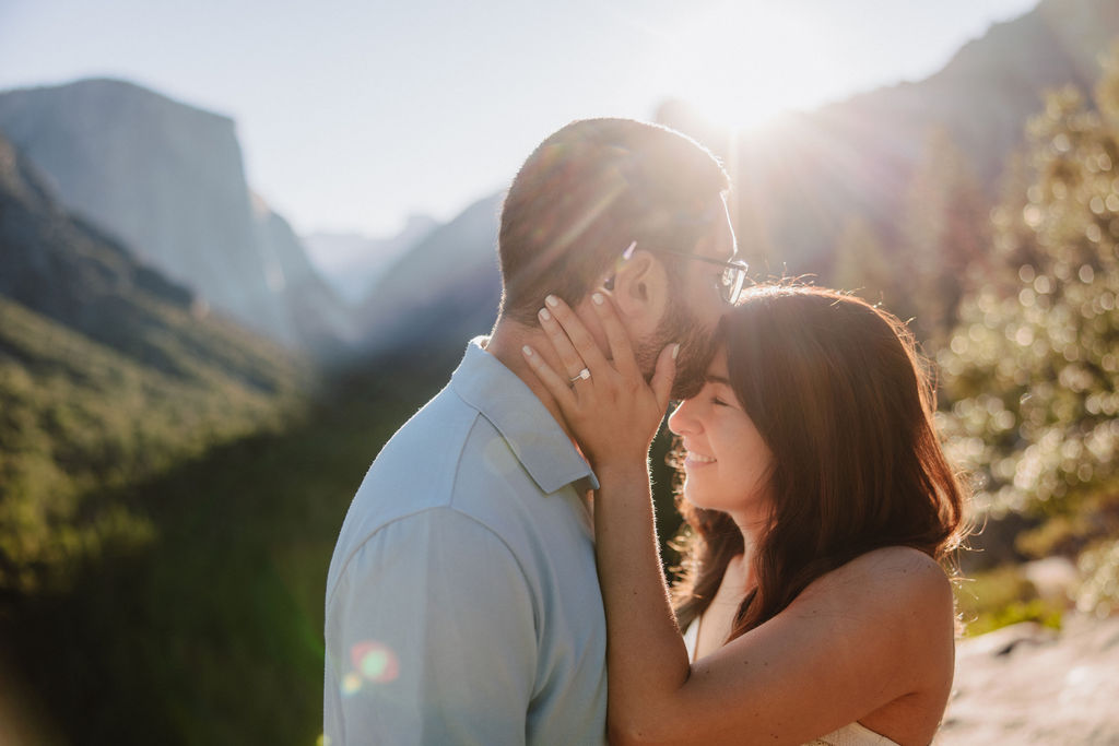 woman holds mans face and smiles while the sun peaks through them for their Yosemite National Park engagement photos