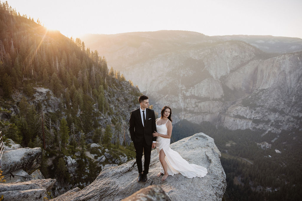 A couple dressed in wedding attire stands on a mountain ledge overlooking a vast rocky landscape with a prominent peak in the background. Best views in yosemite national park