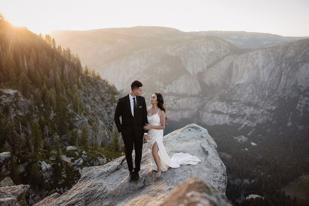 A couple dressed in wedding attire stands on a mountain ledge overlooking a vast rocky landscape with a prominent peak in the background. Best views in yosemite national park