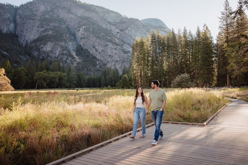 A couple walks on a wooden path through a grassy field with tall trees and mountains in the background for Yosemite national park engagement photos