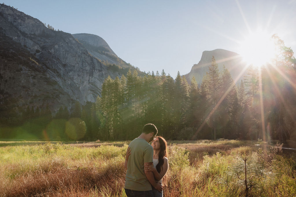 A couple embraces in a sunlit meadow with mountains and tall trees in the background. Sunlight creates lens flare across the scene for their Yosemite national park engagement photos