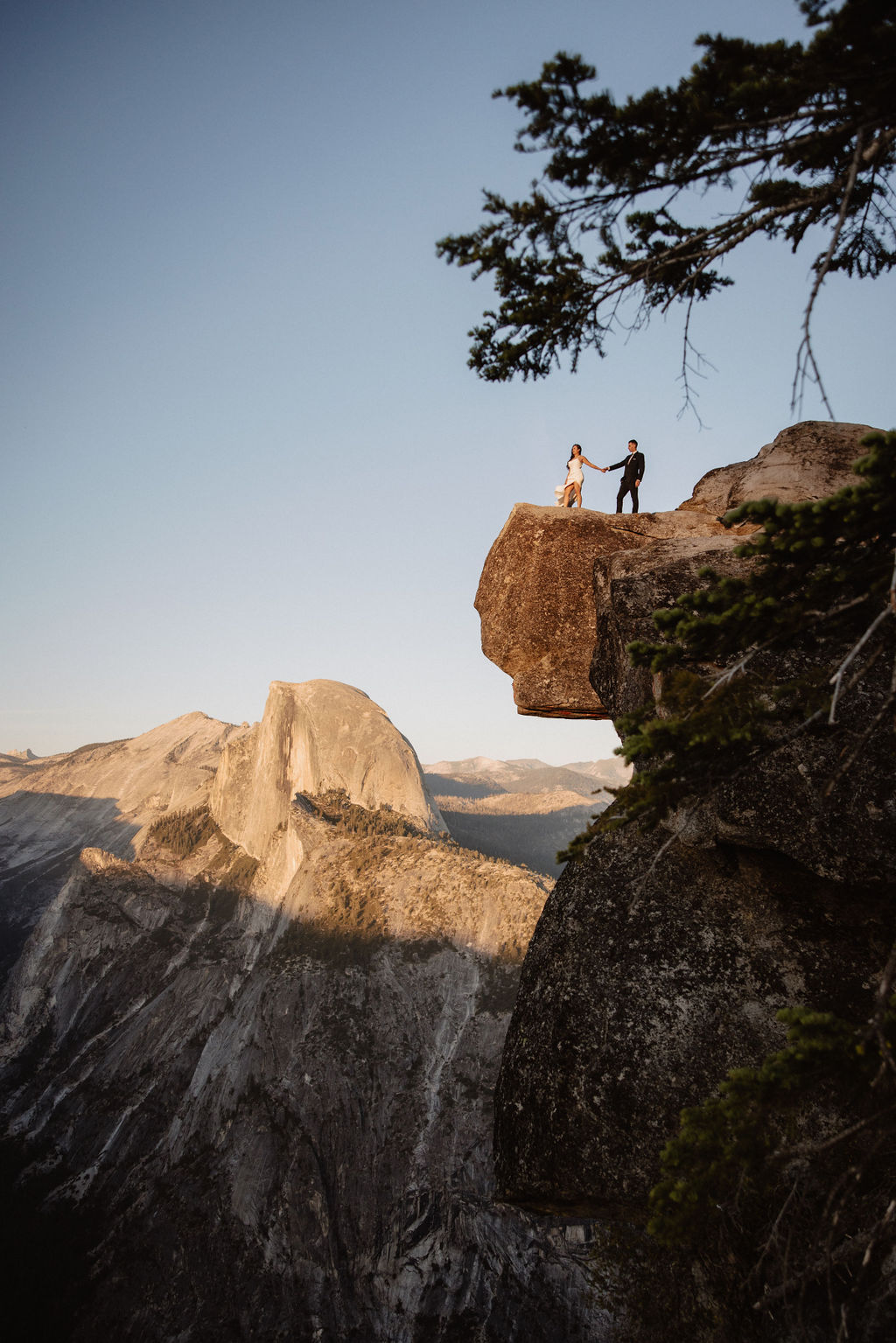 A couple dressed in wedding attire stands on a mountain ledge overlooking a vast rocky landscape with a prominent peak in the background. 