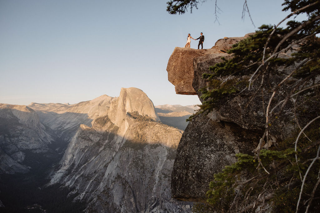 A couple dressed in wedding attire stands on a mountain ledge overlooking a vast rocky landscape with a prominent peak in the background. 