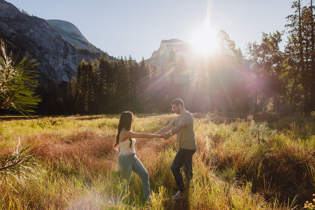 A couple embraces in a sunlit meadow with mountains and tall trees in the background. Sunlight creates lens flare across the scene for their Yosemite national park engagement photos