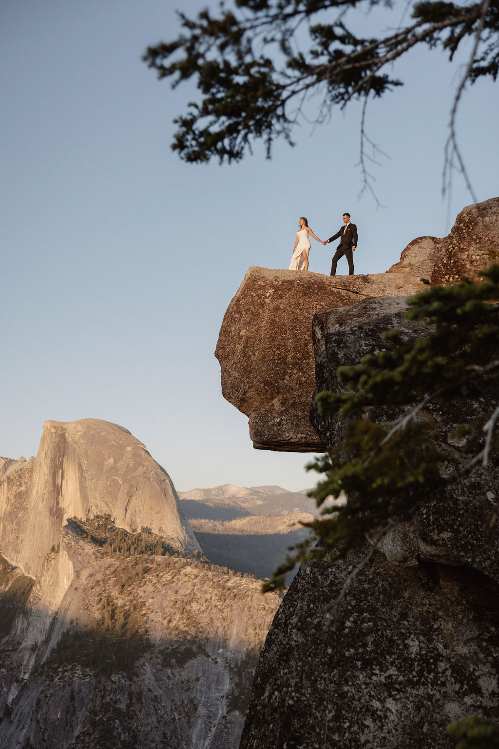  A couple dressed in wedding attire stands on a mountain ledge overlooking a vast rocky landscape with a prominent peak in the background. 