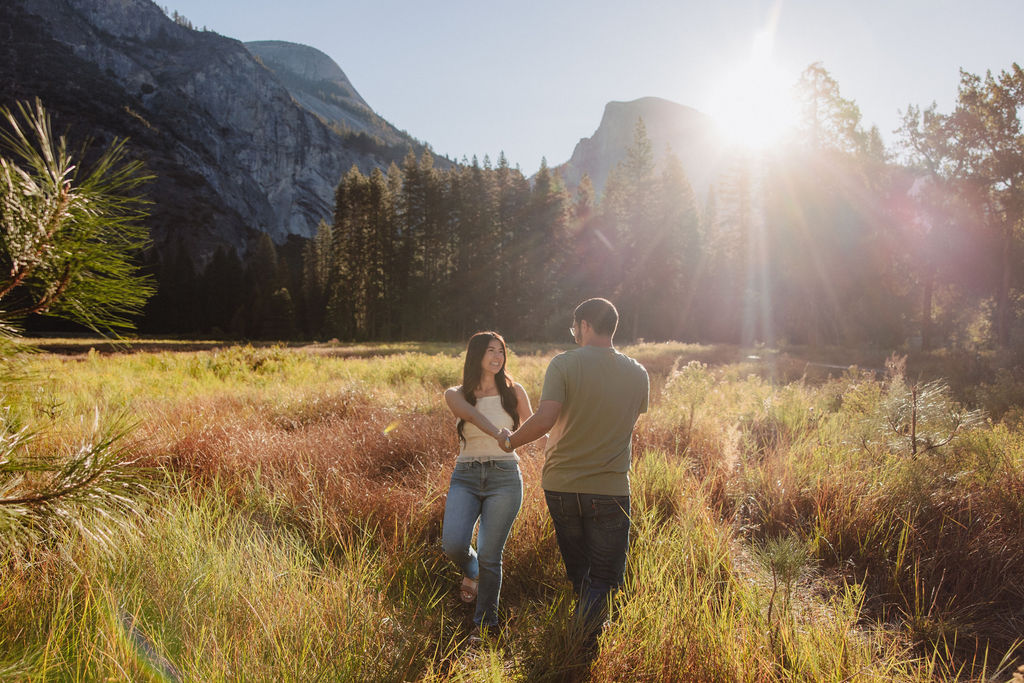 A couple embraces in a sunlit meadow with mountains and tall trees in the background. Sunlight creates lens flare across the scene for their Yosemite national park engagement photos