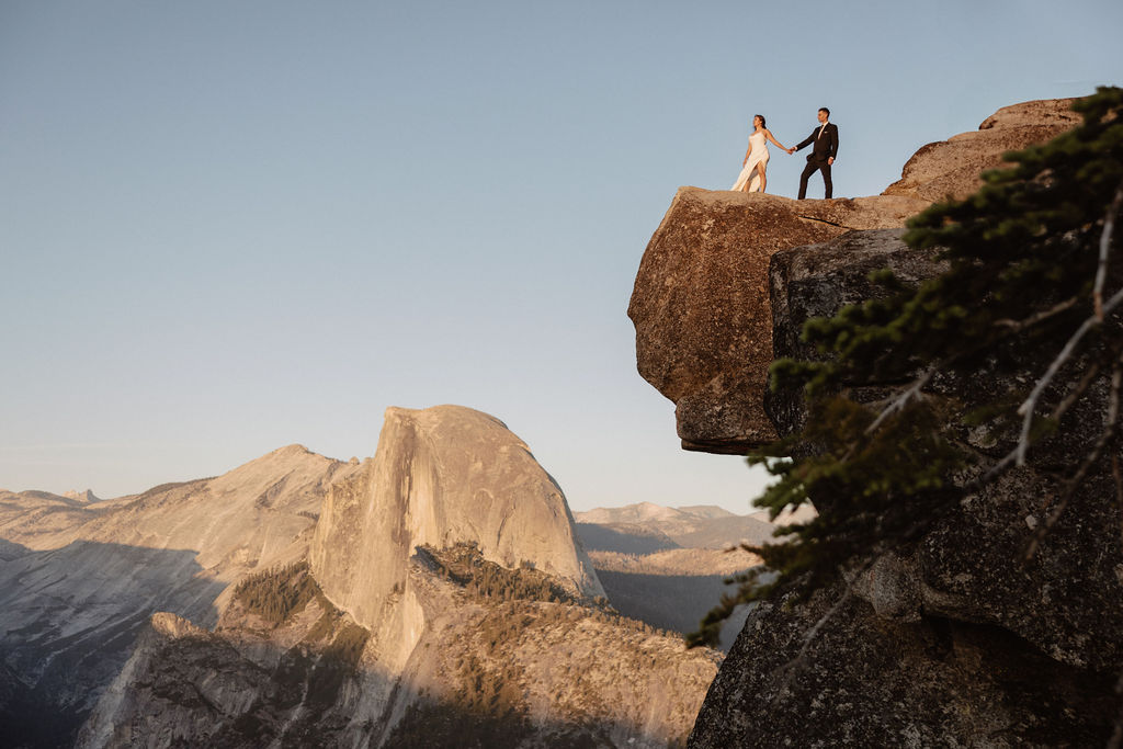 A couple dressed in wedding attire stands on a mountain ledge overlooking a vast rocky landscape with a prominent peak in the background.