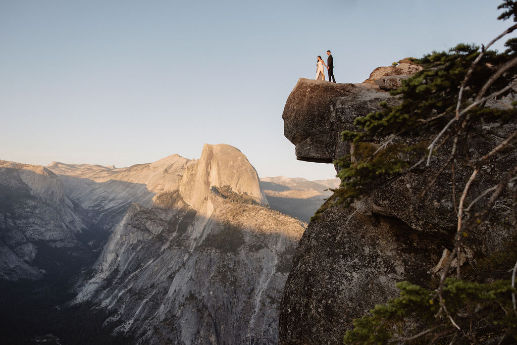 A couple dressed in wedding attire stands on a mountain ledge overlooking a vast rocky landscape with a prominent peak in the background. 