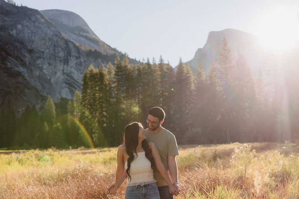 A couple embraces in a sunlit meadow with mountains and tall trees in the background. Sunlight creates lens flare across the scene for their Yosemite national park engagement photos