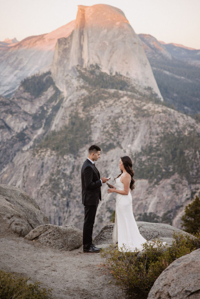 A couple dressed in wedding attire stands on a mountain ledge overlooking a vast rocky landscape with a prominent peak in the background.