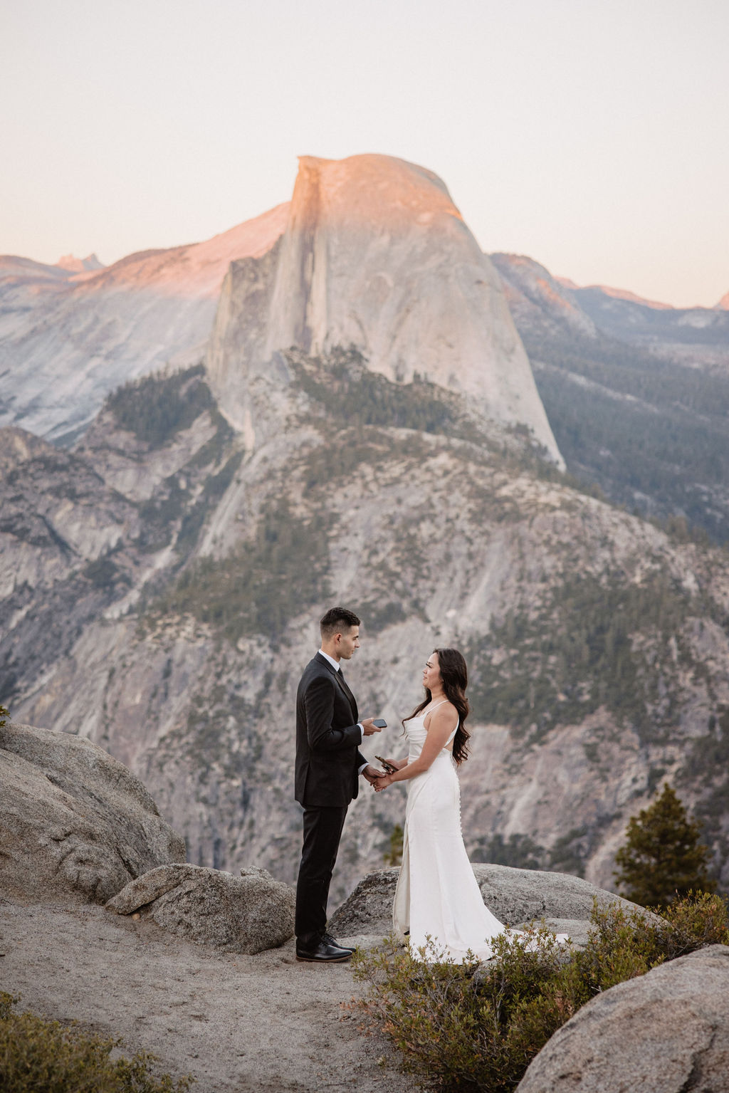 A couple dressed in wedding attire stands on a mountain ledge overlooking a vast rocky landscape with a prominent peak in the background. 