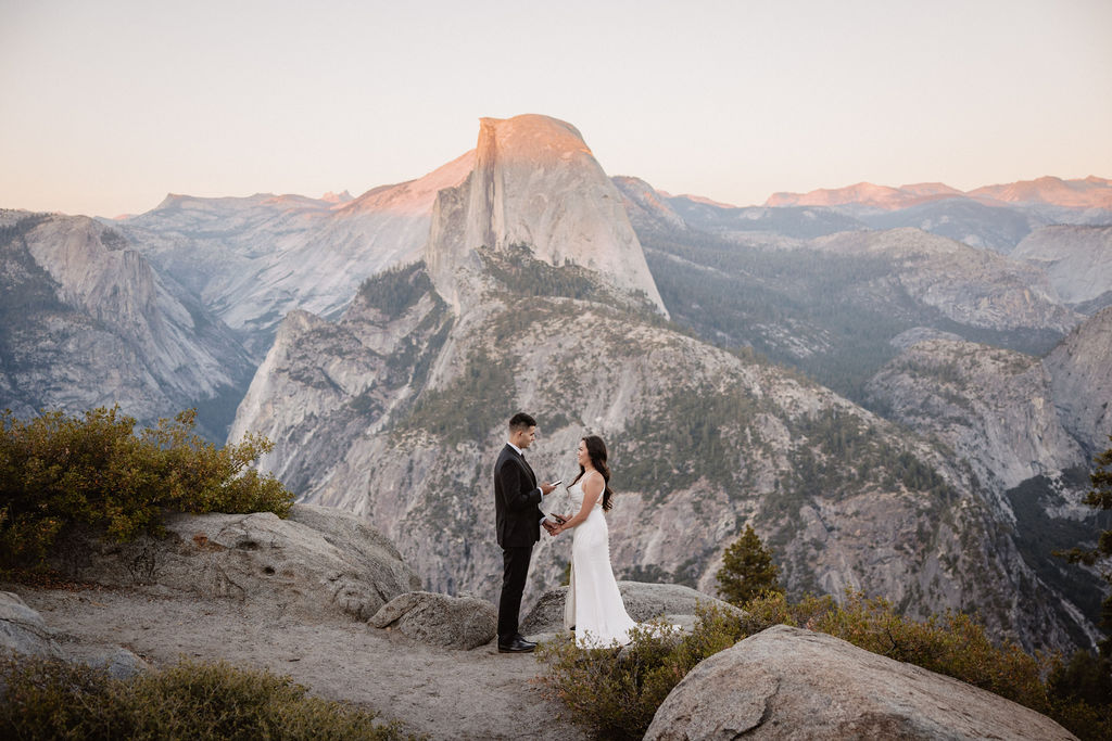 A couple dressed in wedding attire stands on a mountain ledge overlooking a vast rocky landscape with a prominent peak in the background. 