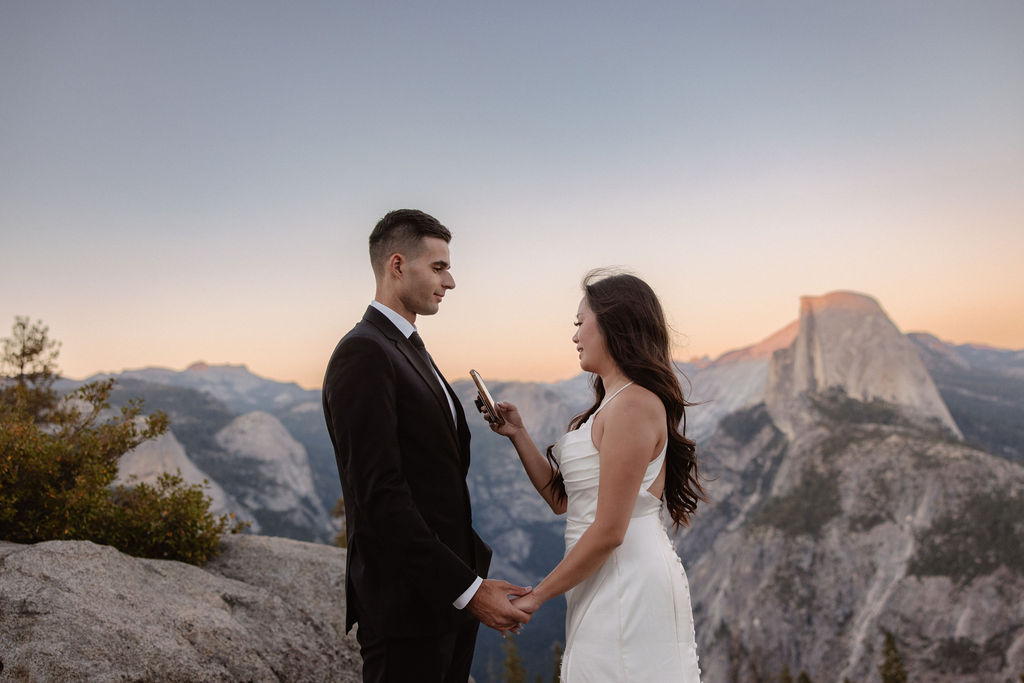 A couple dressed in wedding attire stands on a mountain ledge overlooking a vast rocky landscape with a prominent peak in the background. 