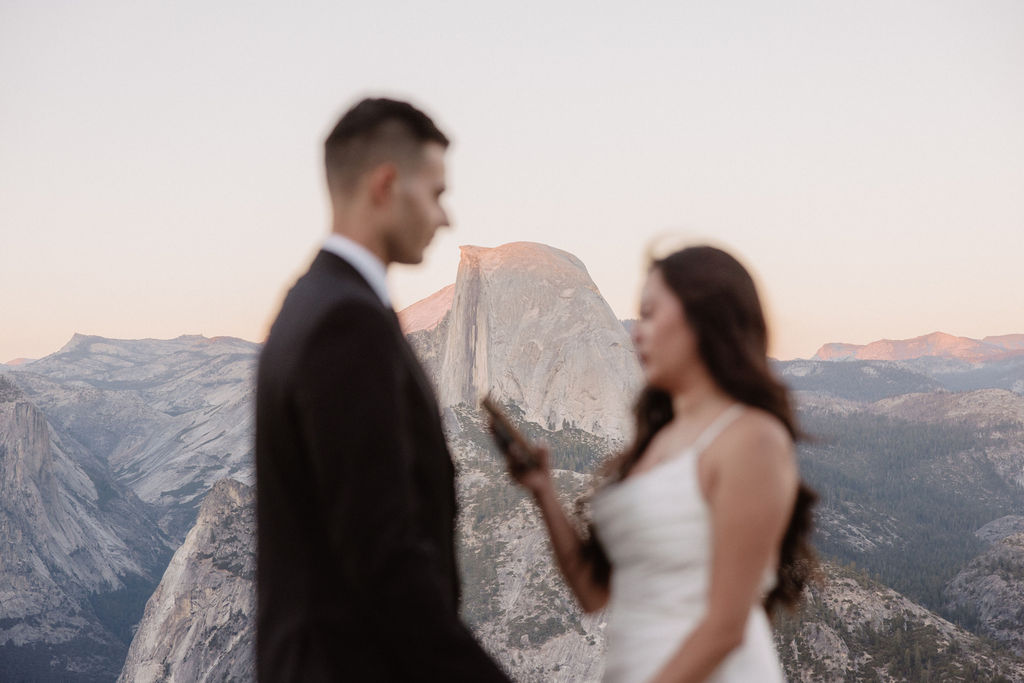 A couple dressed in wedding attire stands on a mountain ledge overlooking a vast rocky landscape with a prominent peak in the background. 