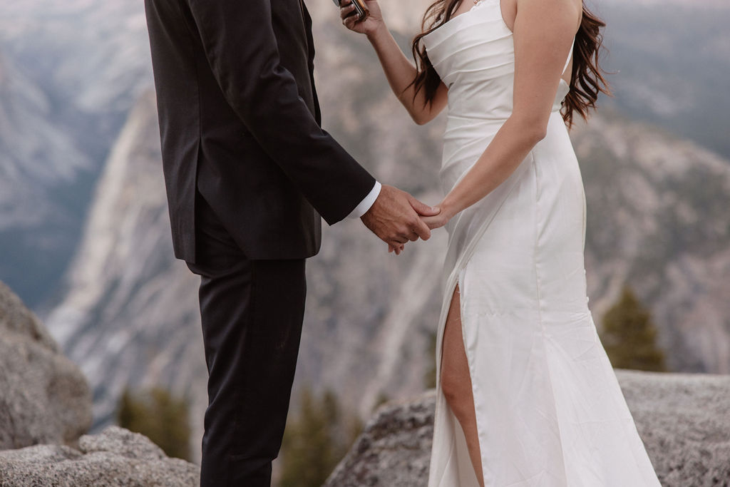 A couple dressed in wedding attire stands on a mountain ledge overlooking a vast rocky landscape with a prominent peak in the background. 