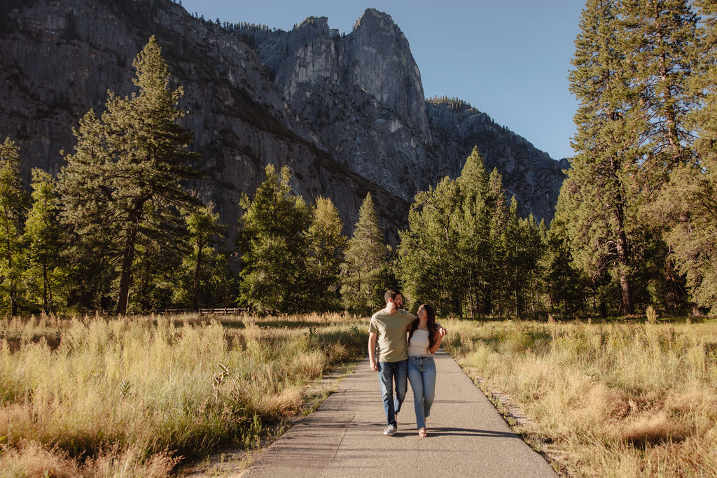 A couple walks on a paved path in a meadow, surrounded by trees and mountains under a clear sky for their yosemite national park engagement photos