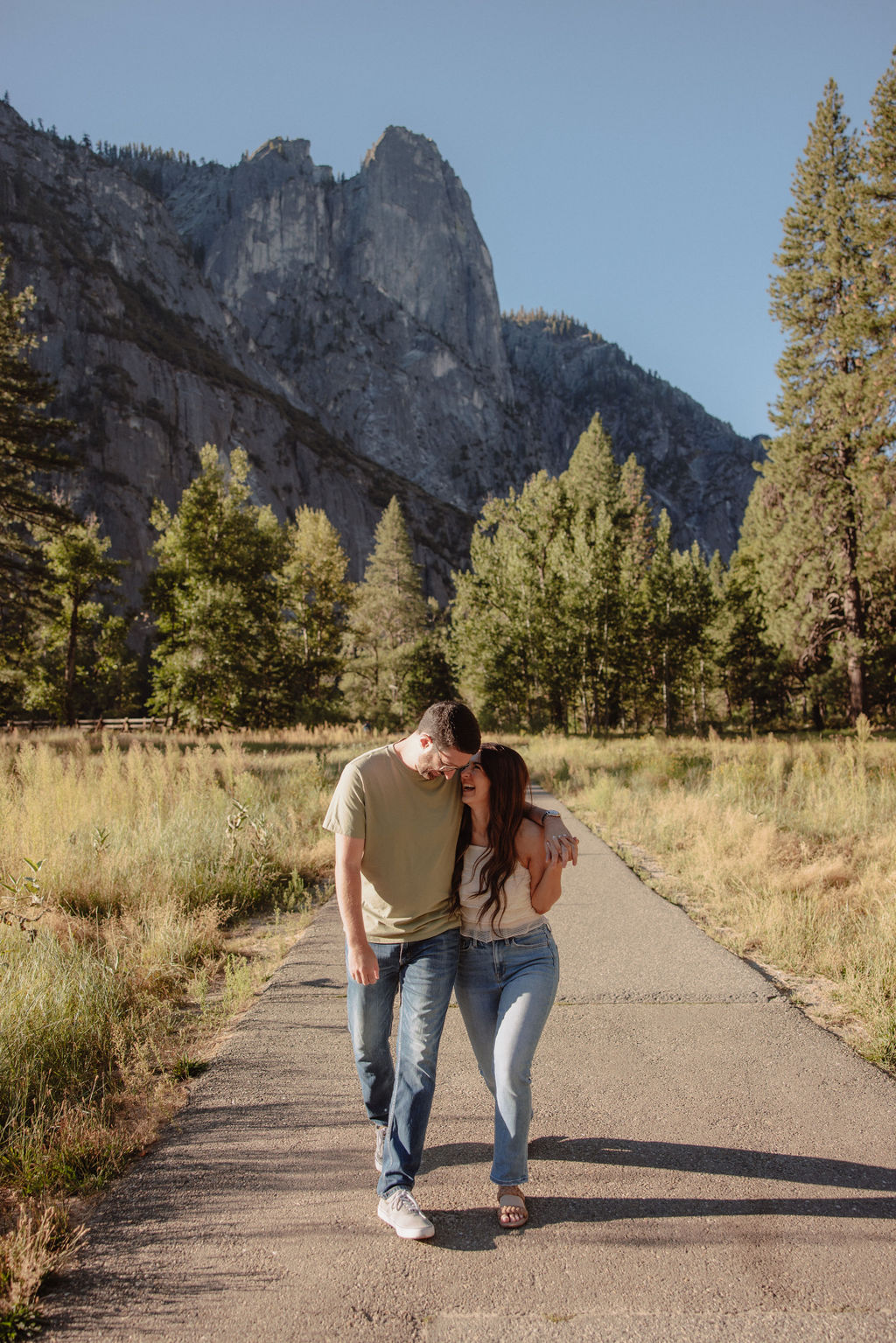 A couple walks arm in arm on a paved path surrounded by trees and mountains under a clear blue sky for their Yosemite National park engagement photos 