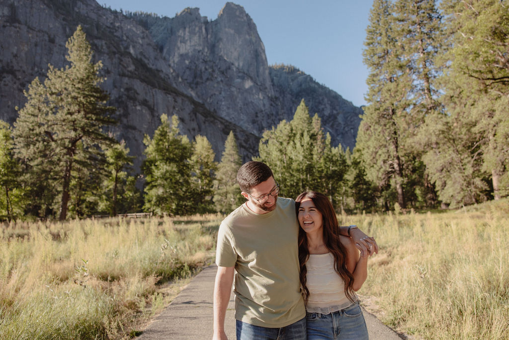 A couple walks on a paved path in a meadow, surrounded by trees and mountains under a clear sky for their yosemite national park engagement photos