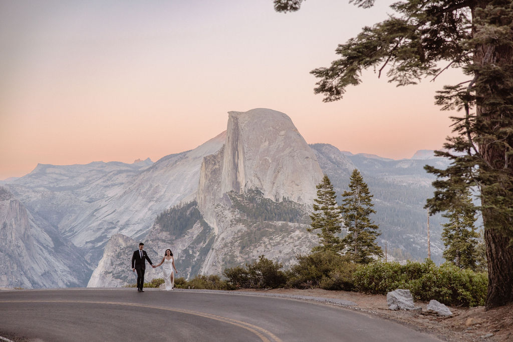 Bride and groom walk hand in hand on a road with Half Dome in Yosemite National Park in the background under a pastel sky.