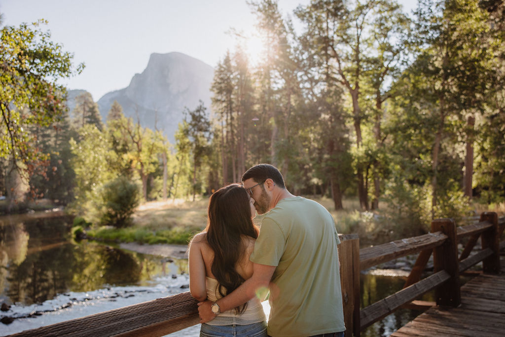 A couple stands on a bridge overlooking a serene river, surrounded by trees, with a mountain in the background under a sunny sky.