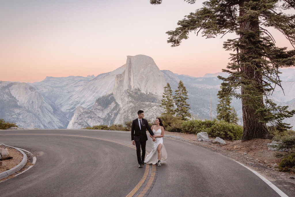 Bride and groom walk hand in hand on a road with Half Dome in Yosemite National Park in the background under a pastel sky.