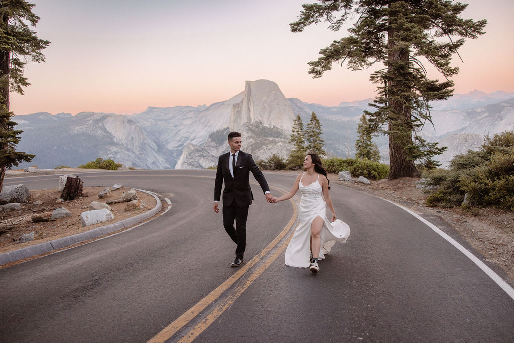 Bride and groom walk hand in hand on a road with Half Dome in Yosemite National Park in the background under a pastel sky.