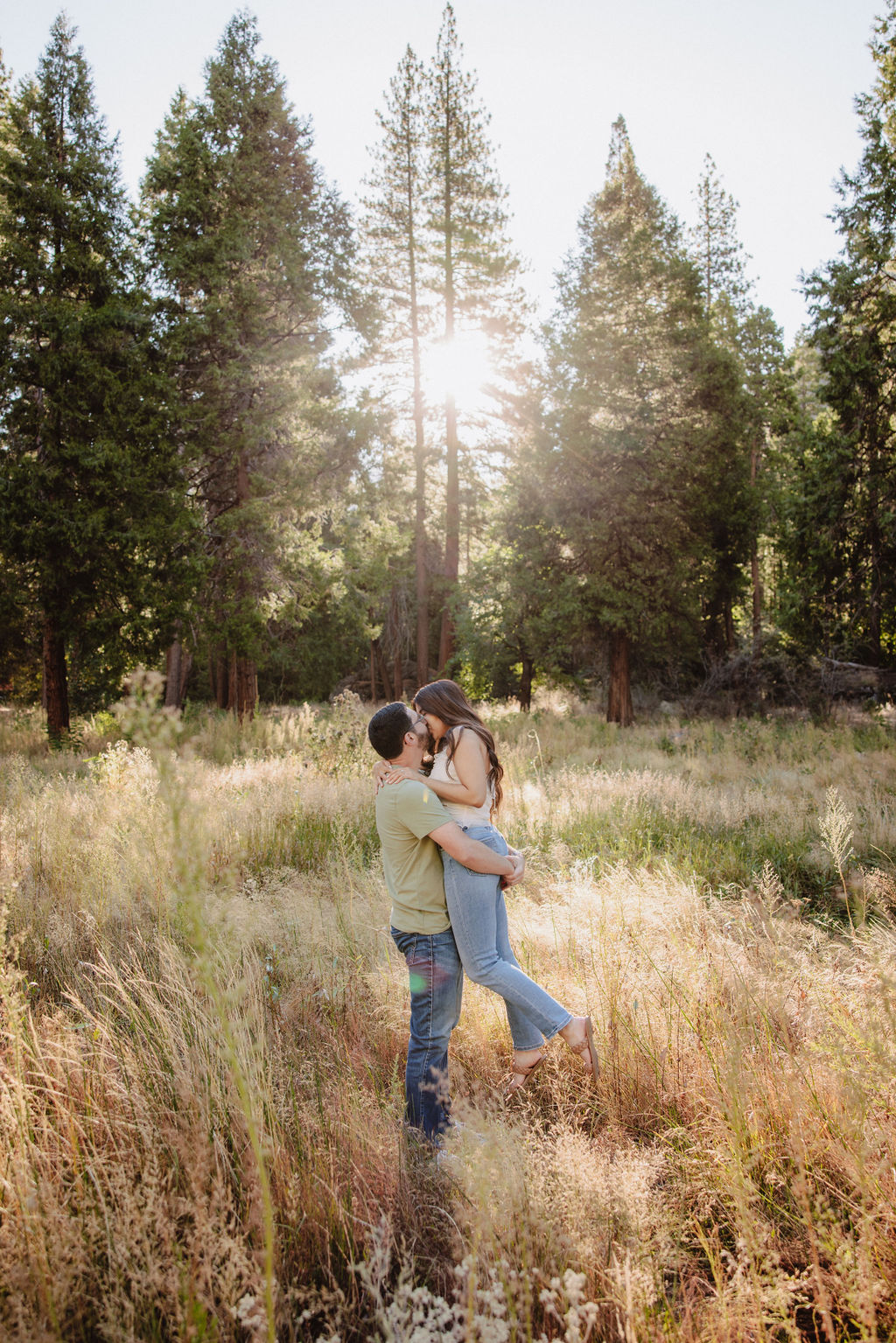 A couple embraces in a sunlit forest clearing with tall grass and trees in the background.