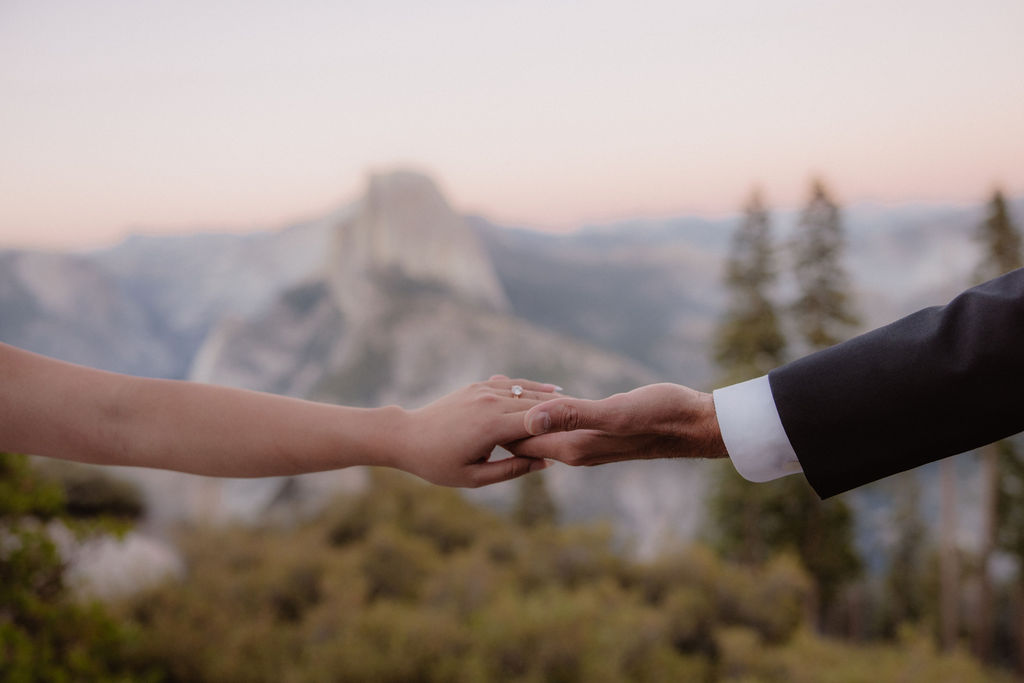 A couple dressed in wedding attire stands on a mountain ledge overlooking a vast rocky landscape with a prominent peak in the background. 