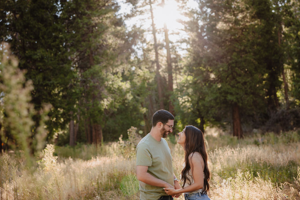 A couple embraces in a sunlit forest clearing with tall grass and trees in the background.