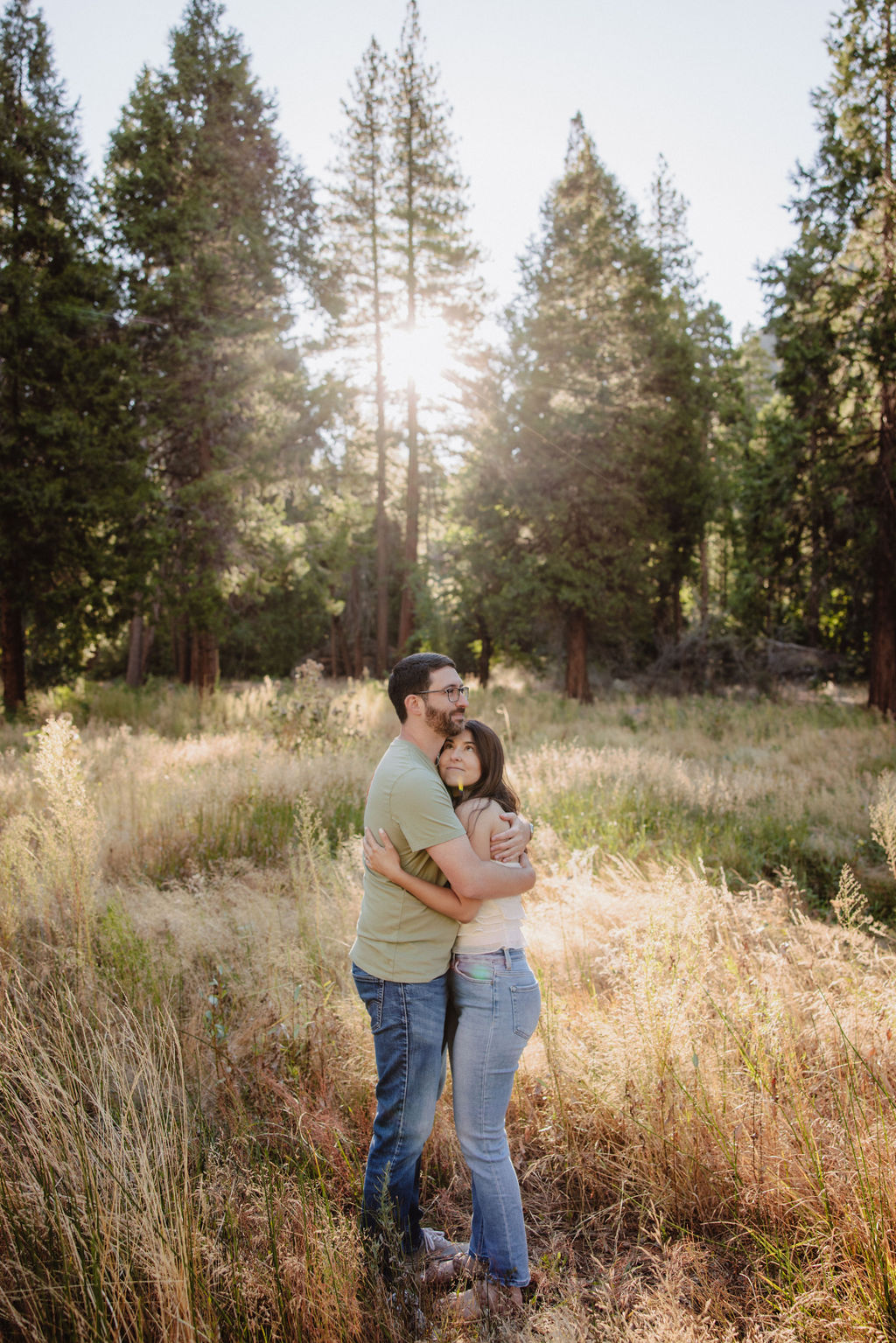 A couple embraces in a sunlit forest clearing with tall grass and trees in the background.