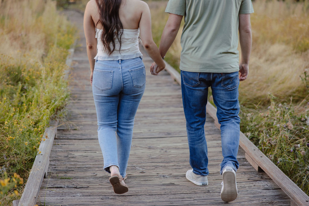 Two people in casual clothing walk hand in hand on a wooden path surrounded by grass.