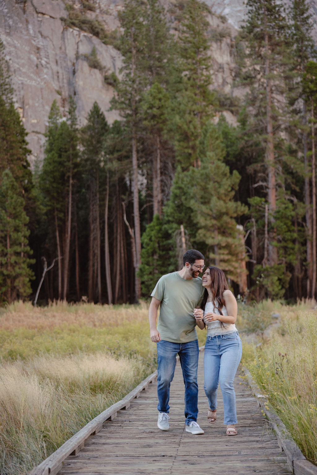 A couple embraces in a sunlit forest clearing with tall grass and trees in the background.