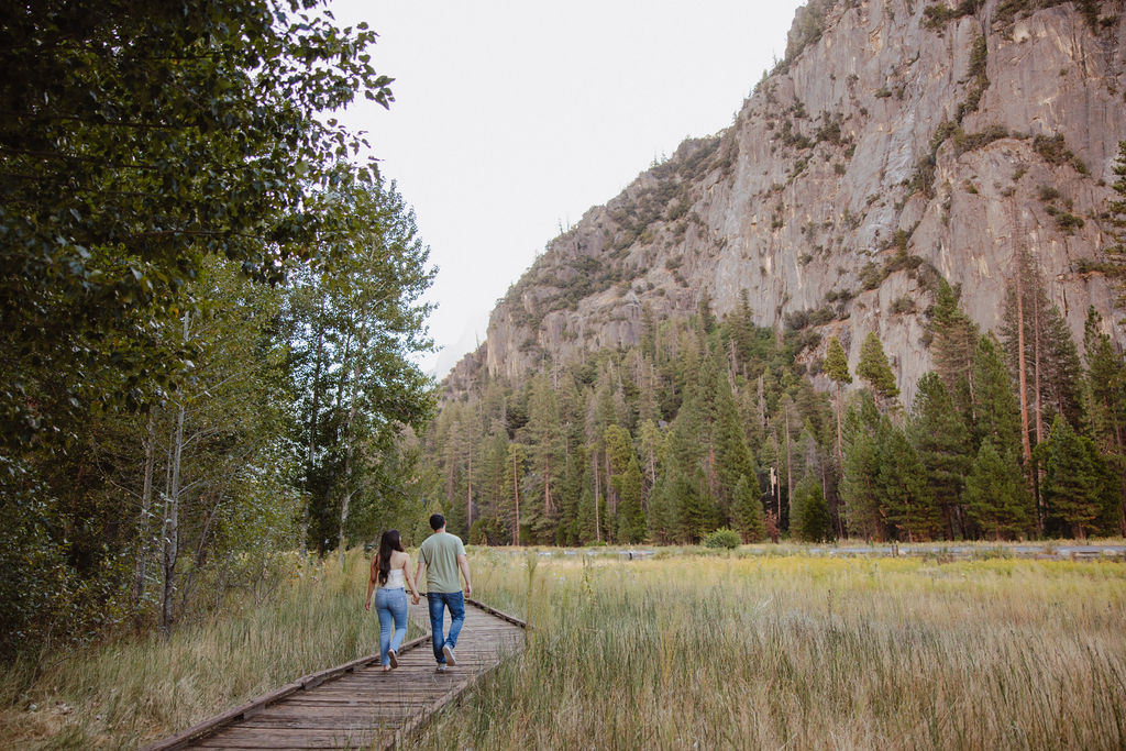 A couple walks along a wooden path through a grassy field, with trees and a rocky cliff in the background for their yosemite national park engagement photos