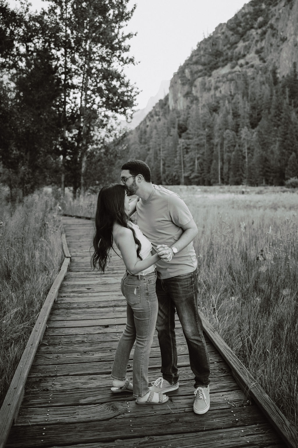 A couple walks along a wooden path through a grassy field, with trees and a rocky cliff in the background for their yosemite national park engagement photos