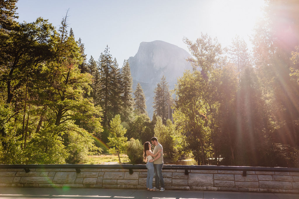 A couple walks along a wooden path through a grassy field, with trees and a rocky cliff in the background for their yosemite national park engagement photos