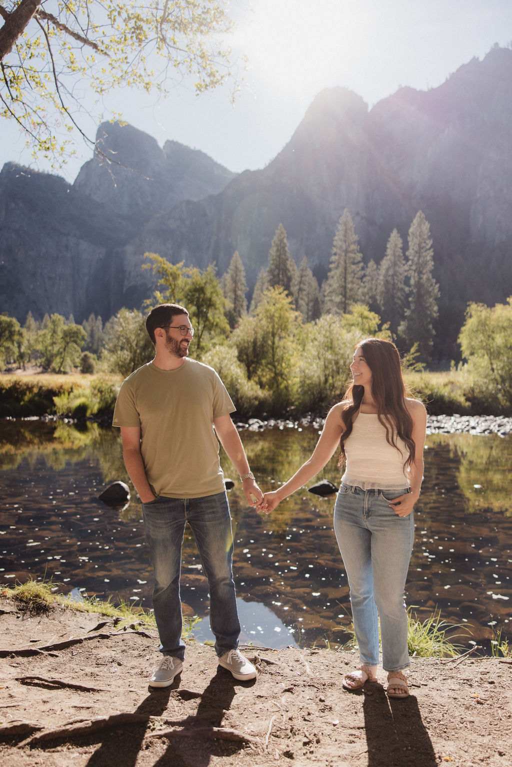 A couple holds hands by a river with trees and mountains in the background under a clear sky for their yosemite national park engagement photos