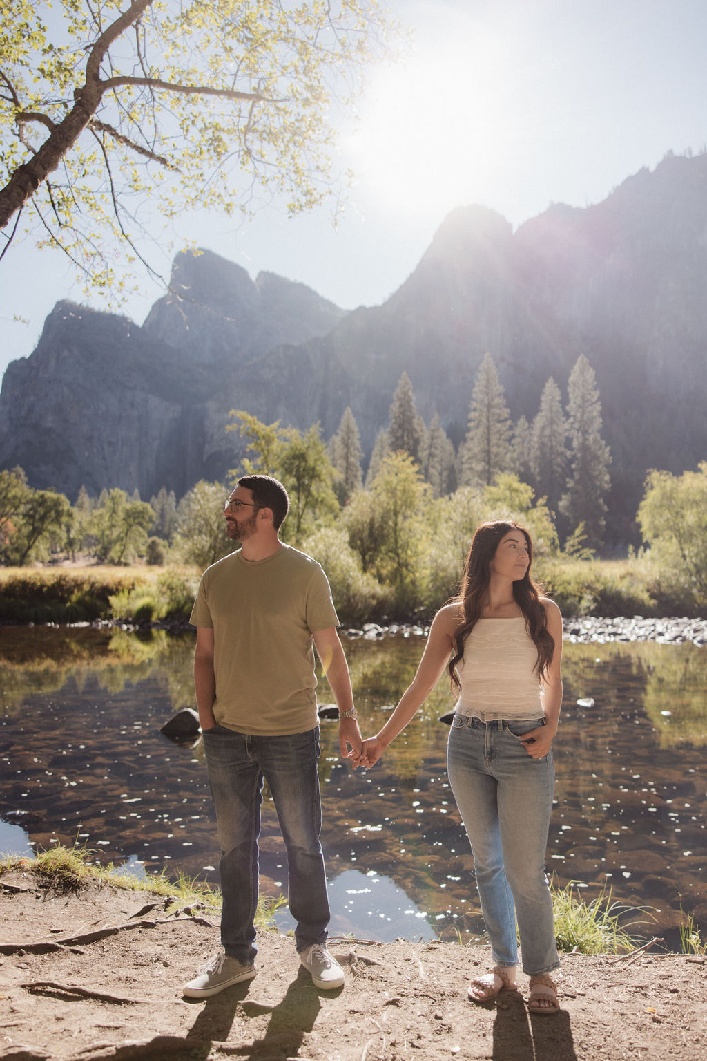 A couple holds hands by a river with trees and mountains in the background under a clear sky for their yosemite national park engagement photos