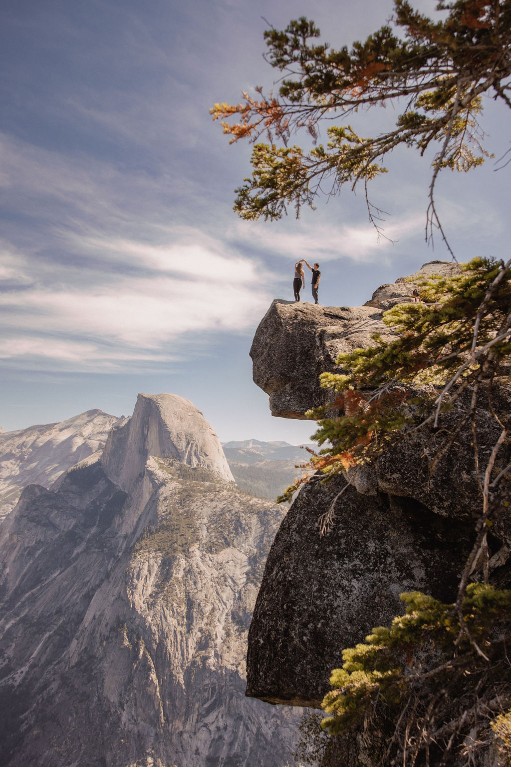 Two people stand on a rocky cliff edge with a mountain landscape in the background, under a partly cloudy sky. | How to Plan a Surprise Proposal at Yosemite National Park