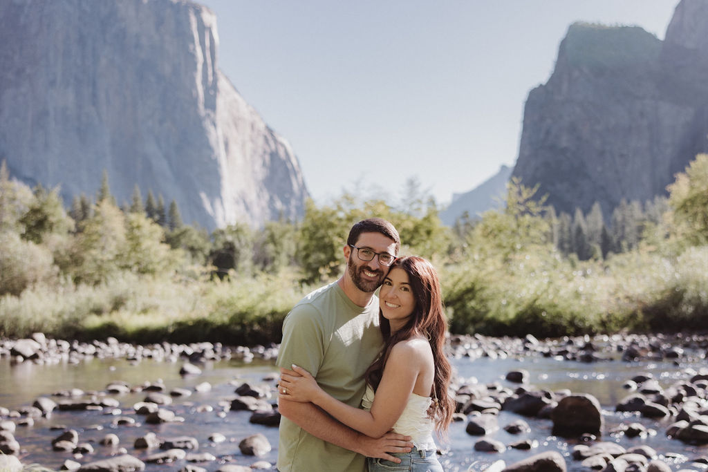 A couple smiling and embracing in front of a rocky river with tall mountains and trees in the background 