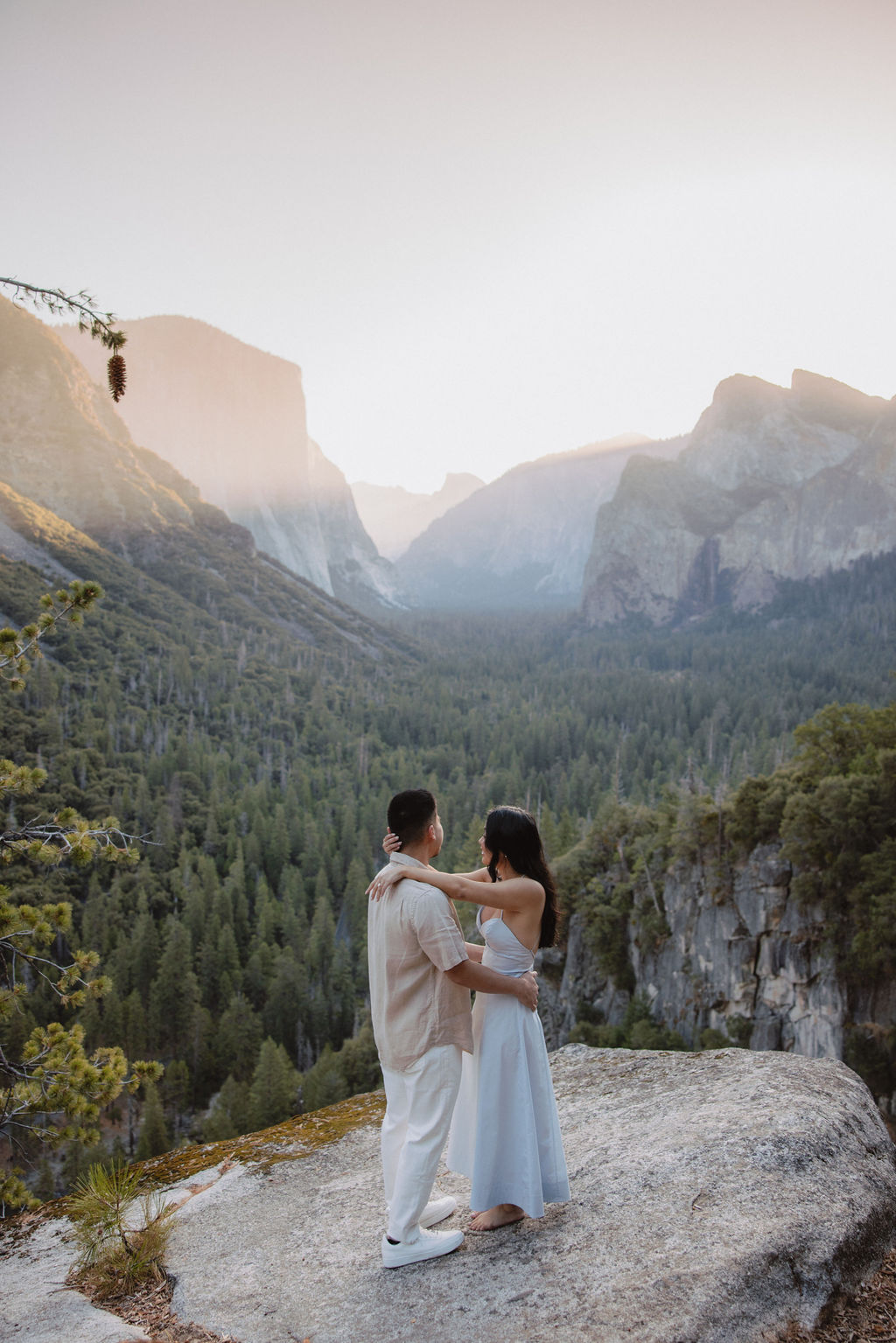 A couple embraces on a rocky ledge overlooking a forested valley with mountains in the background at sunset. | How to Plan a Surprise Proposal at Yosemite National Park