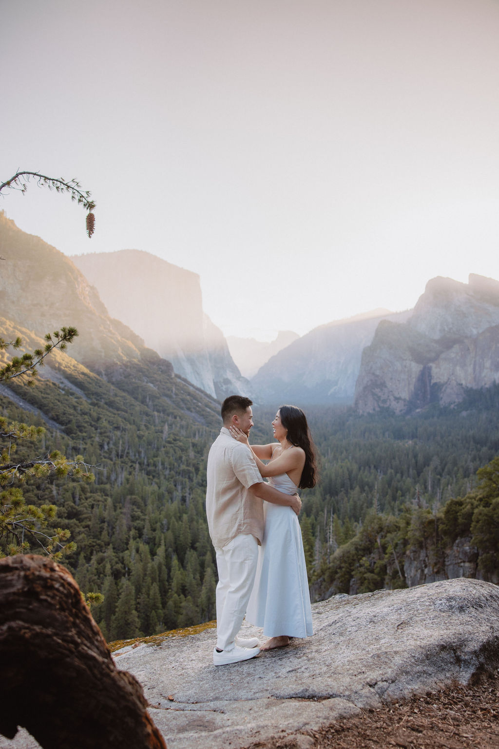 A couple embraces on a rocky ledge overlooking a forested valley with mountains in the background at sunset. | How to Plan a Surprise Proposal at Yosemite National Park