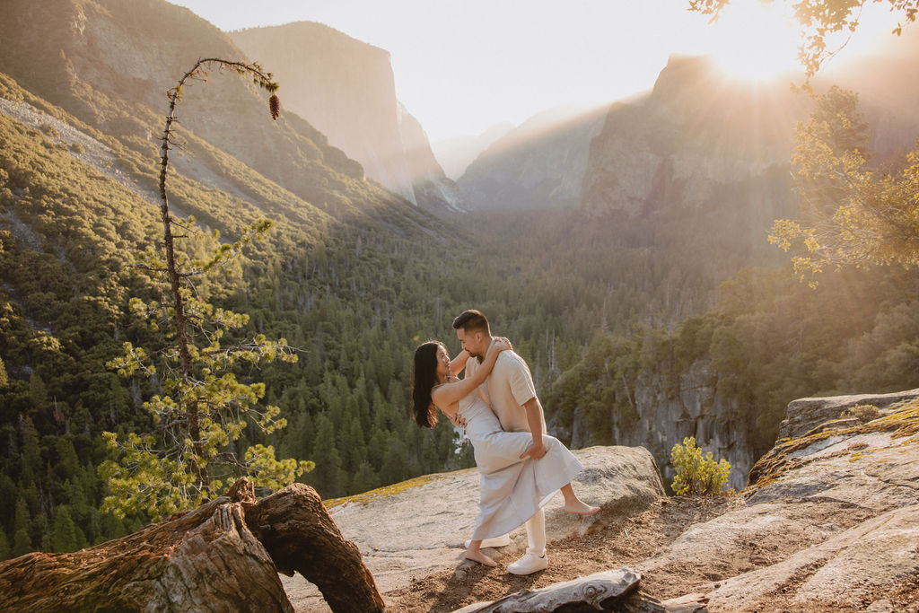 A couple embraces on a rocky ledge overlooking a forested valley with mountains in the background at sunset. | How to Plan a Surprise Proposal at Yosemite National Park